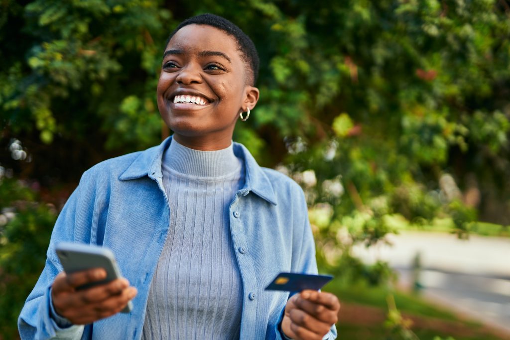 Young african american woman smiling happy holding smartphone and credit card at the city