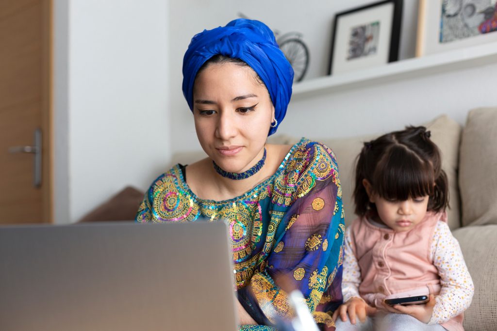 Young Muslim woman using laptop while her young daughter is watc