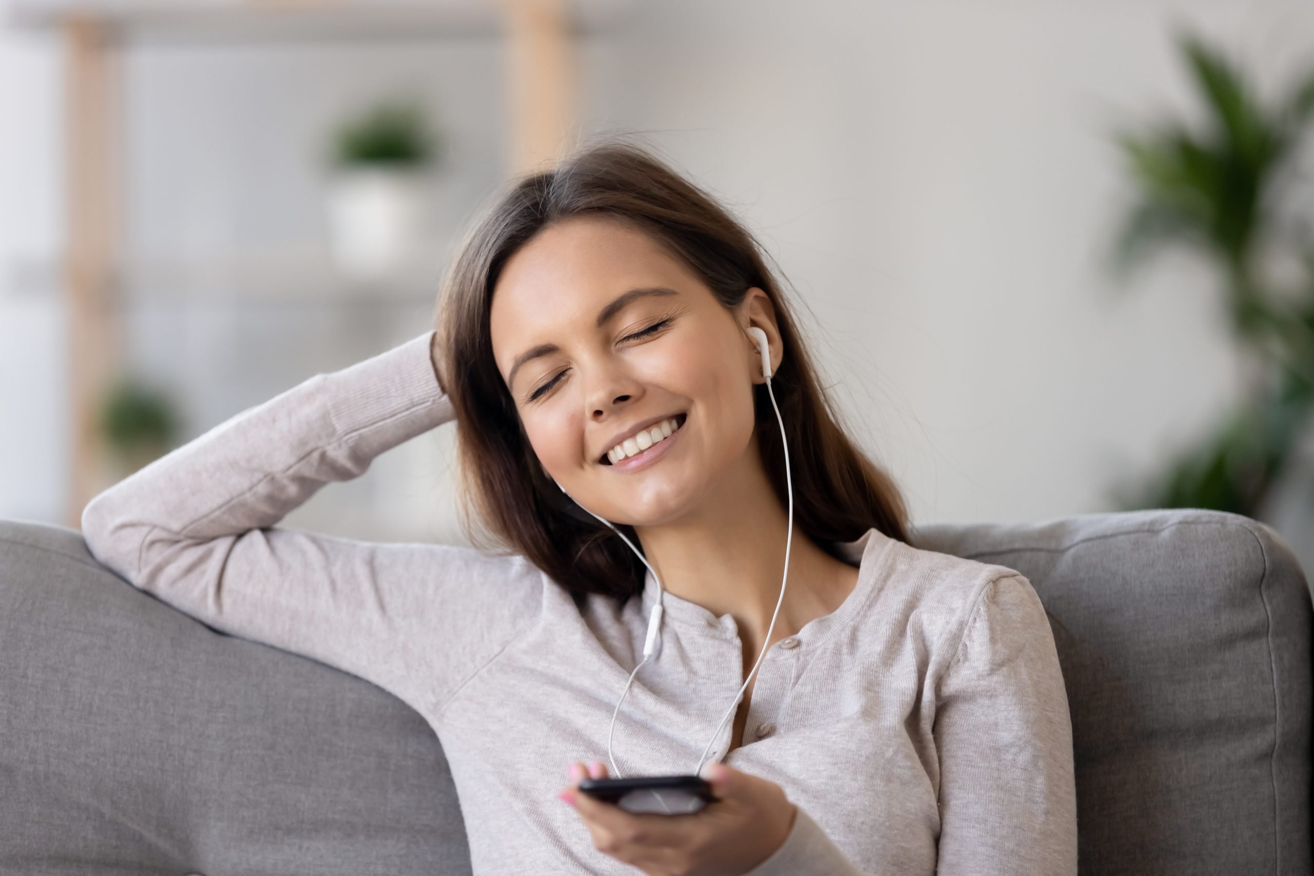 Woman wearing earphones sitting on couch holding phone listening music