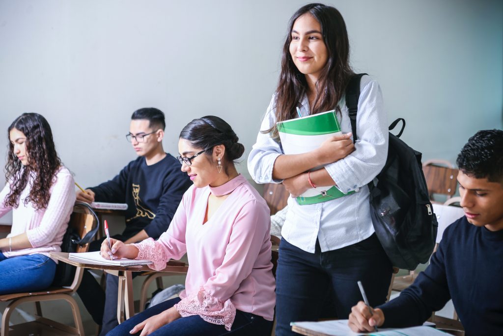 White female student holding a white notebook while standing in class.