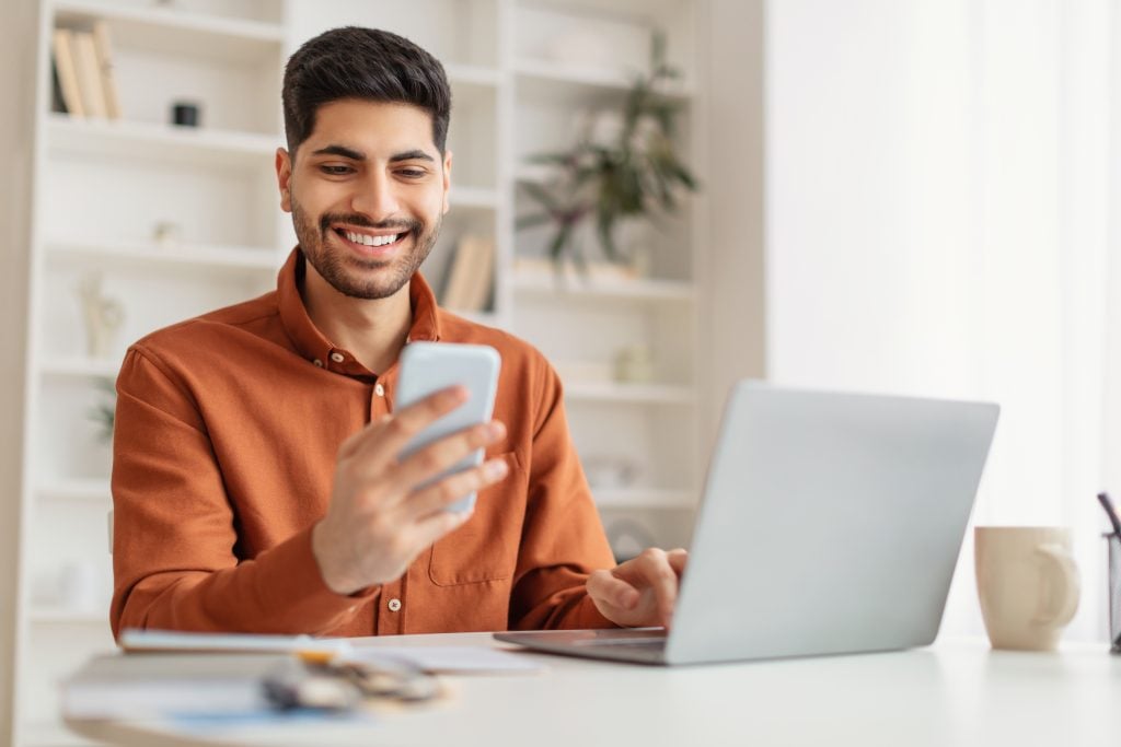 Business And Telecommuting Concept. Portrait of smiling young Arabic man using smartphone and modern laptop computer, reading text message sitting at desk with gadgets, working remotely at home office