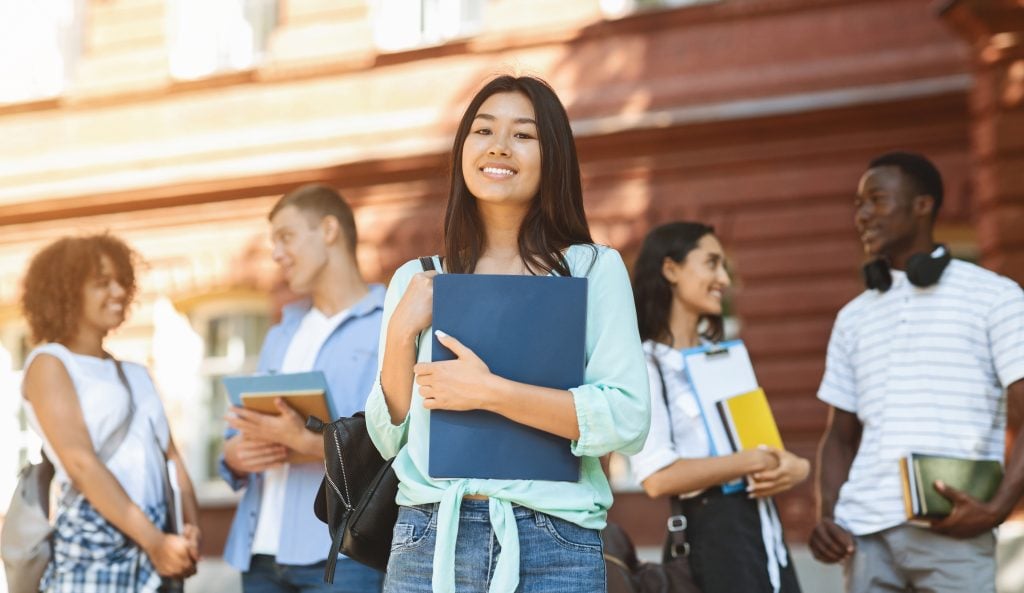 Loans For Education Concept. Portrait Of Happy Asian Student Girl Posing Outdoors In Campus With Her Classmates And University Building On Background