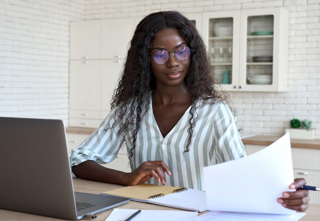 Young intelligent African black businesswoman in glasses sitting at desk reading bank loan legal financial documents using pc laptop computer at modern home office. Distant remote work concept.