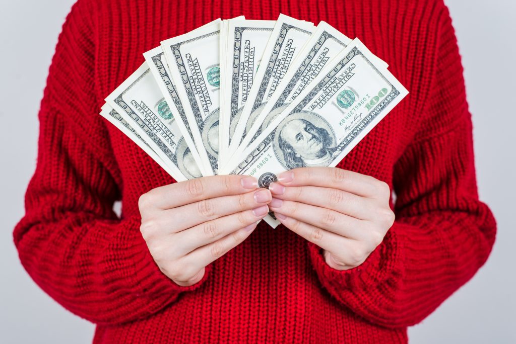 Cropped close up photo of person in bright knitted jumper holding collection of dollars isolated grey background