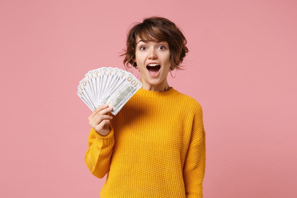 Shocked young brunette woman girl in yellow sweater posing isolated on pastel pink background studio portait. People lifestyle concept. Mock up copy space. Hold fan of cash money in dollar banknotes