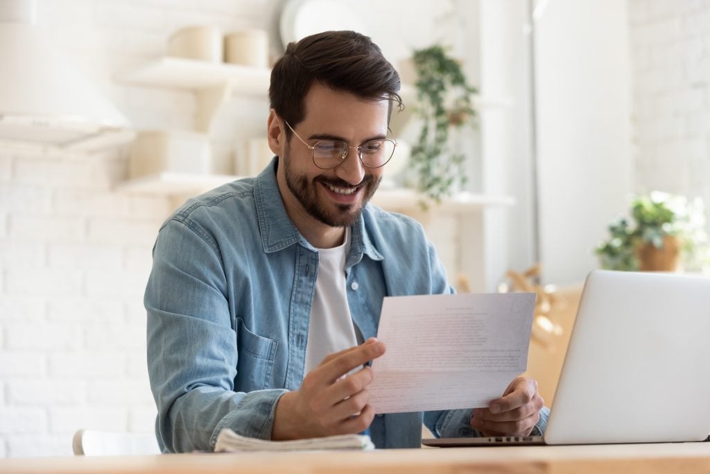 Smiling young man reading good news in paper notification, sitting at table with laptop, happy satisfied male wearing glasses holding document, job promotion, loan approval, great exam result