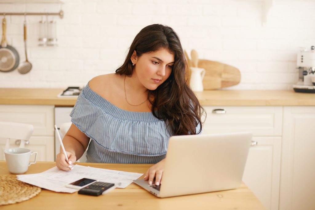 Serious young chubby female sitting in kitchen with portable computer, filling in papers looking at screen. Focused plump housewife making online payments via internet using laptop and calculator