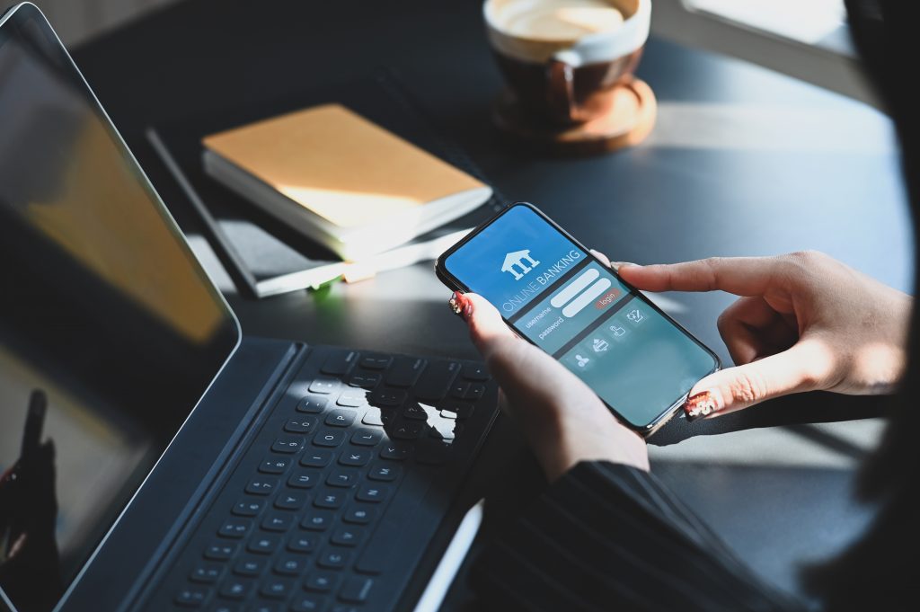 Cropped shot of executive woman holding smartphone in hands while doing online financial transaction by her smartphone in front computer tablet at the modern working desk.