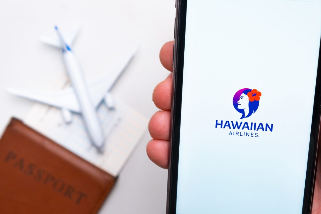 Hawaiian airline company logo on the screen of mobile phone in man hand on the background of passport, boarding pass and plane, September 2021, San Francisco, USA.
