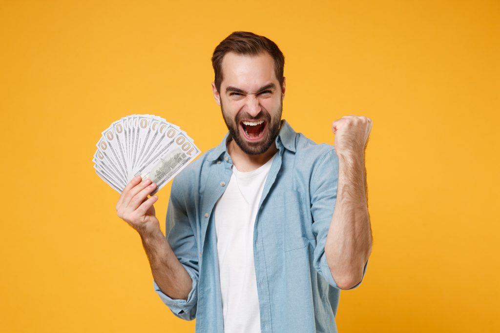 Crazy young man in blue shirt posing isolated on yellow orange background. People lifestyle concept. Mock up copy space. Holding fan of cash money in dollar banknotes, doing winner gesture, screaming