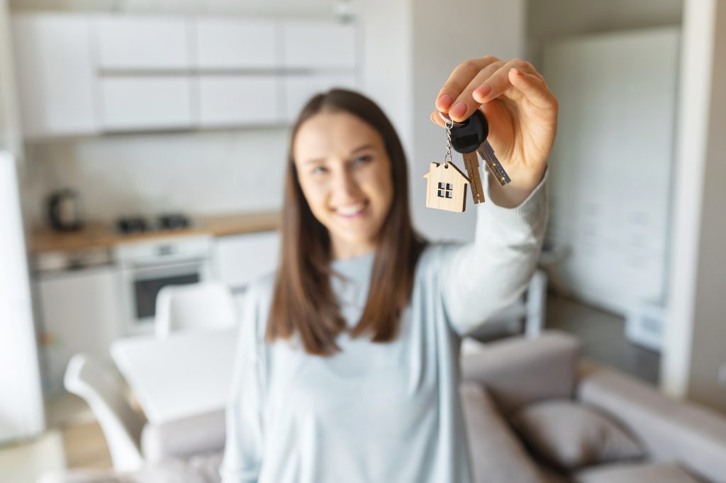 Selective focus on the key in hand of happy young woman, cheerful female moved in new house, rent apartment, bought own real estate, the key with keyring in shape of little house