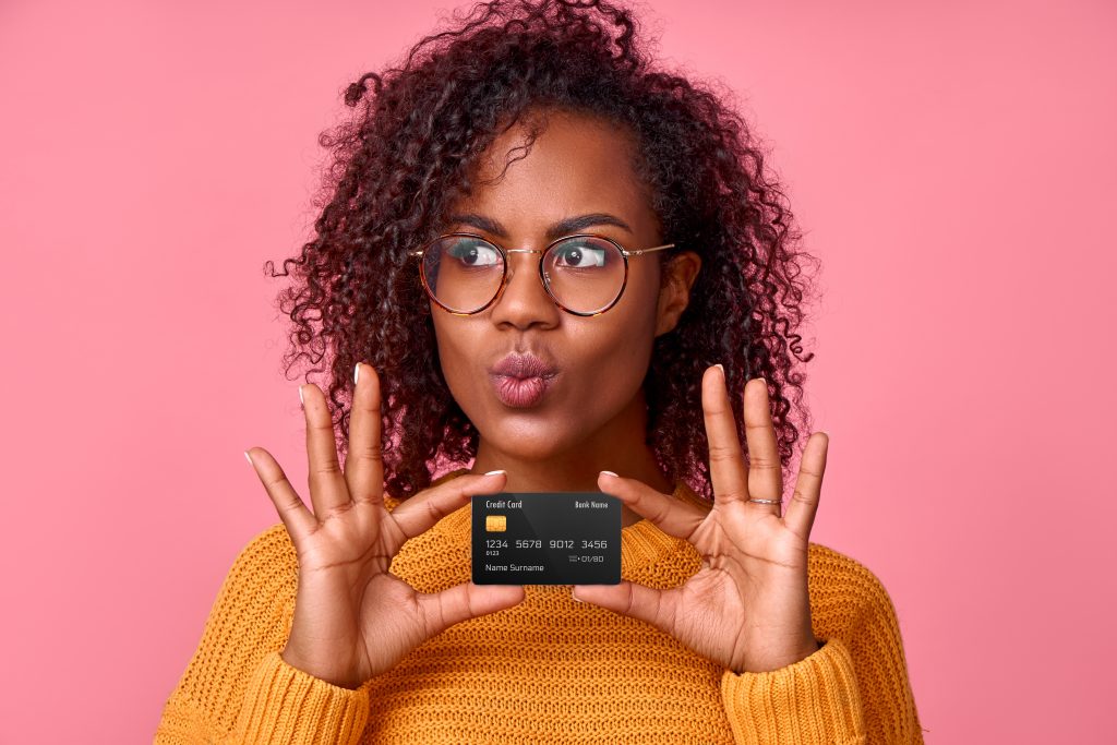 Portrait of young pretty african american woman depicting kiss holding bank credit card in hands, going to buy something in an online store, over pink studio background. Place for advertising