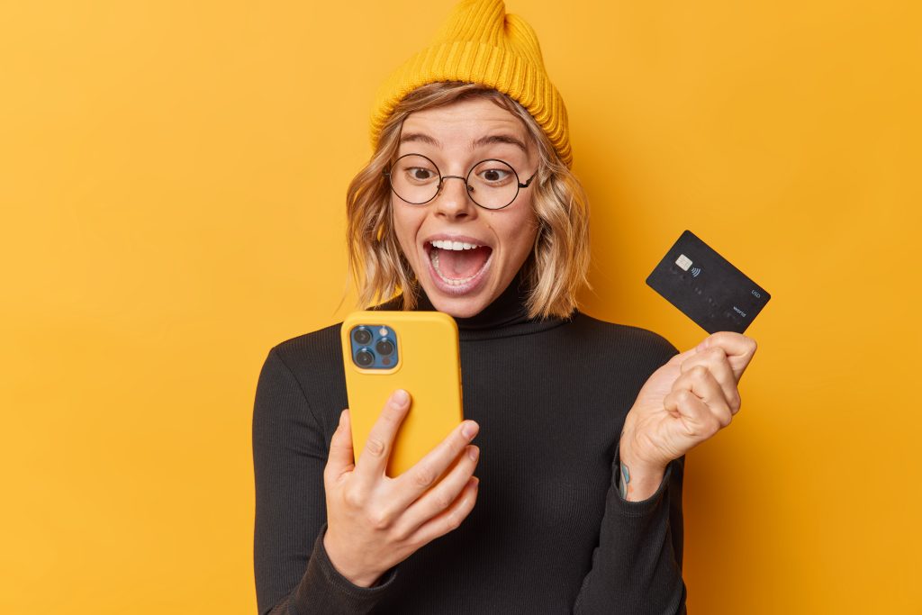 Surpised happy woman wears hat and casual black turtleneck looks at smartphone app screen holds credit card uses bank account for paying online isolated over yellow background gets cashback.