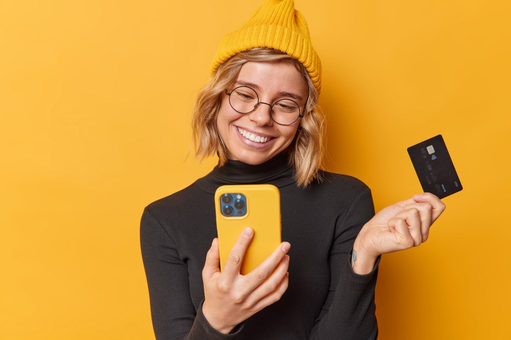 Online payment and purchasing concept. Glad young woman uses smartphone for paying in internet for purchase holds banking card books ticket on plane makes shopping poses indoor against yellow wall