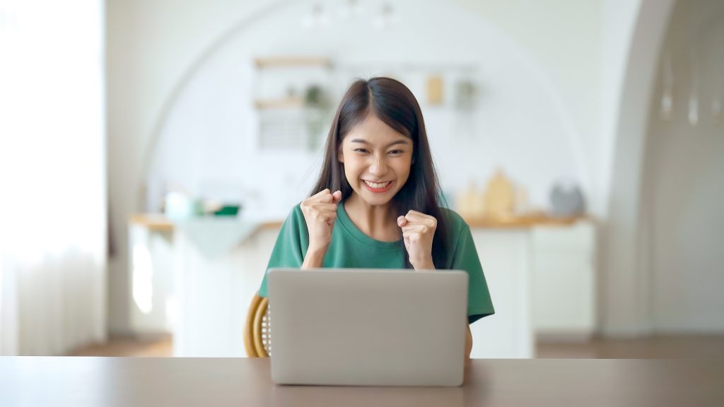 Funny euphoric young asian woman celebrating winning or getting ecommerce shopping offer on computer laptop. Excited happy girl winner looking at notebook celebrating success