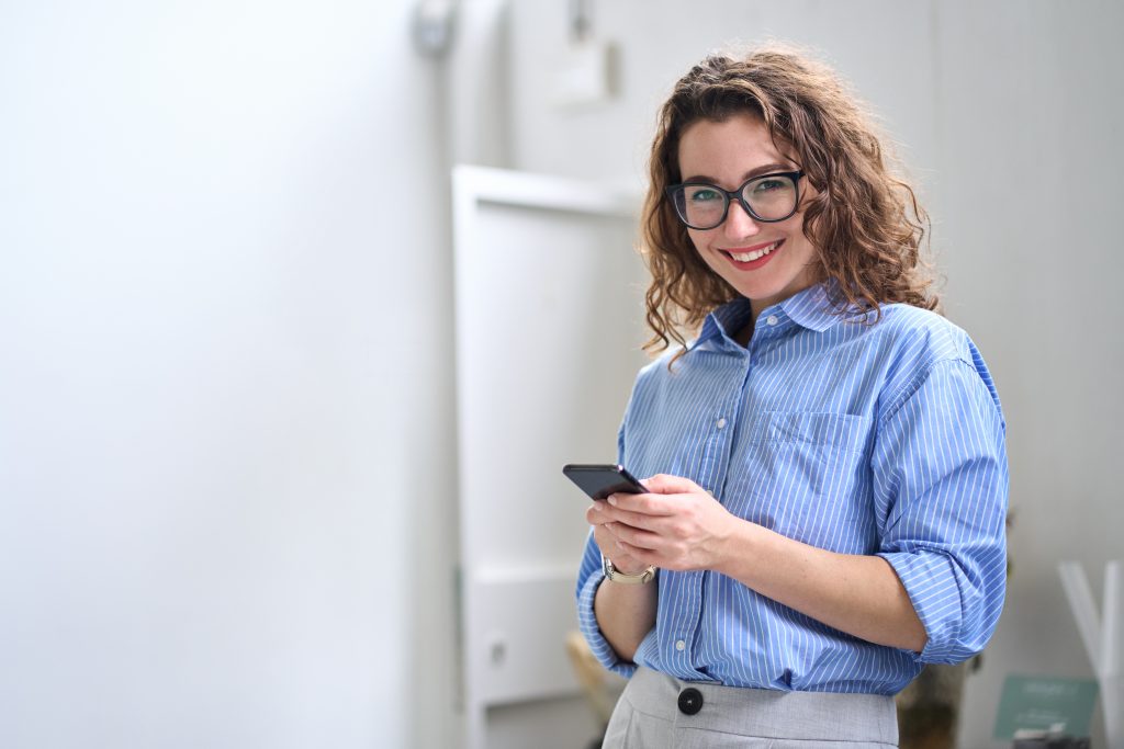 Happy young business woman using cellphone standing in office using mobile.