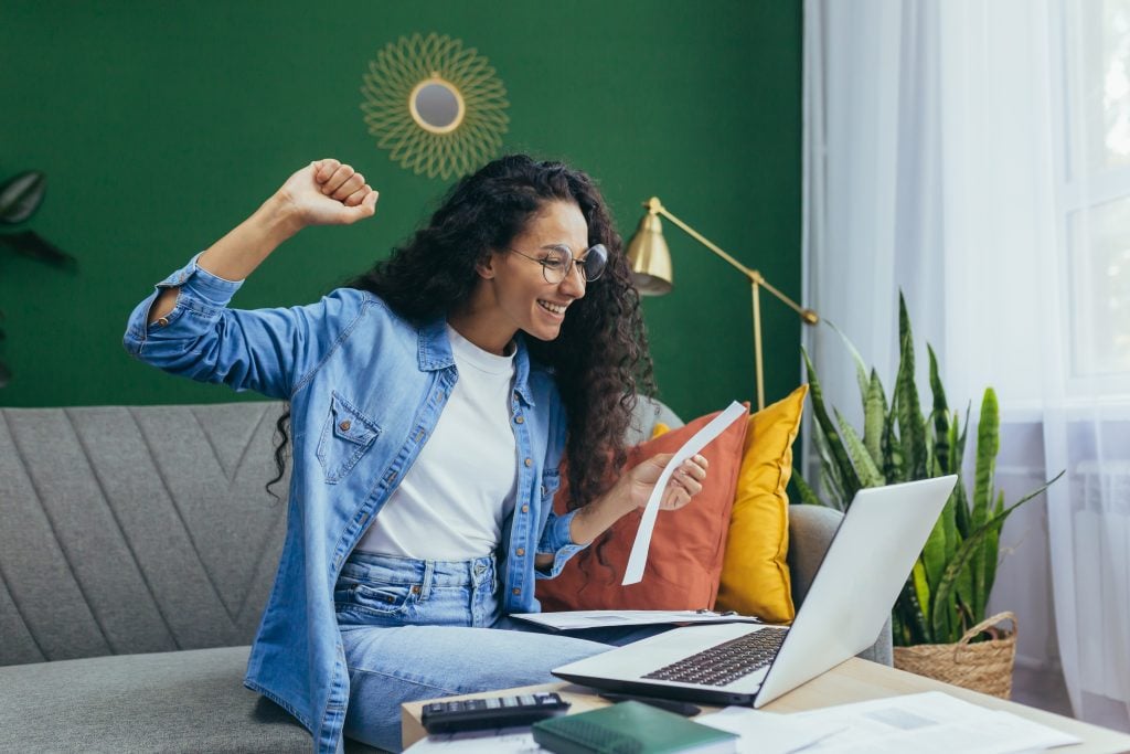Happy hispanic woman at home doing paperwork and calculating household budget, sitting on couch in living room and using laptop at work, holding hand up victory and triumph gesture