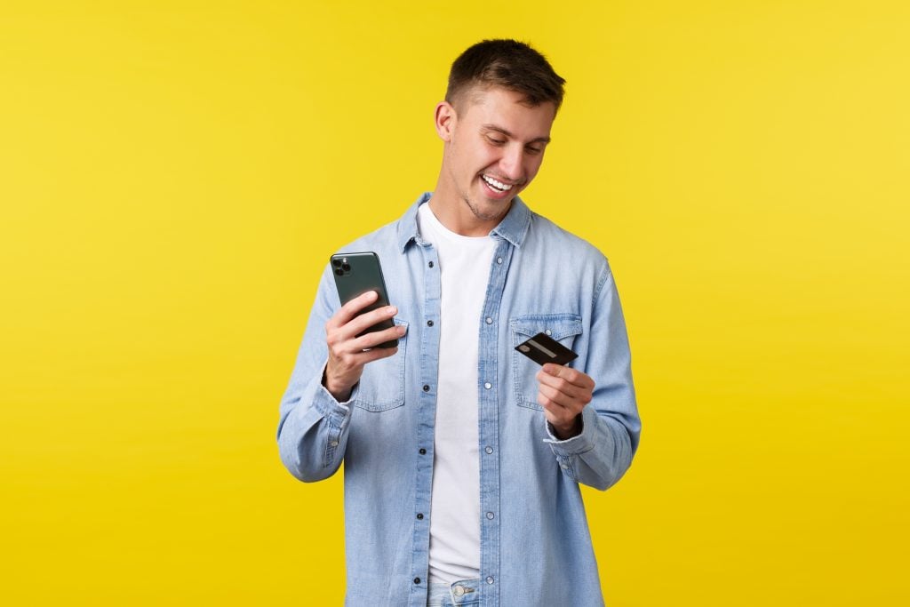 Technology, lifestyle and advertisement concept. Handsome happy man making online order, booking flight tickets with smartphone app, looking at credit card and holding mobile phone, yellow background