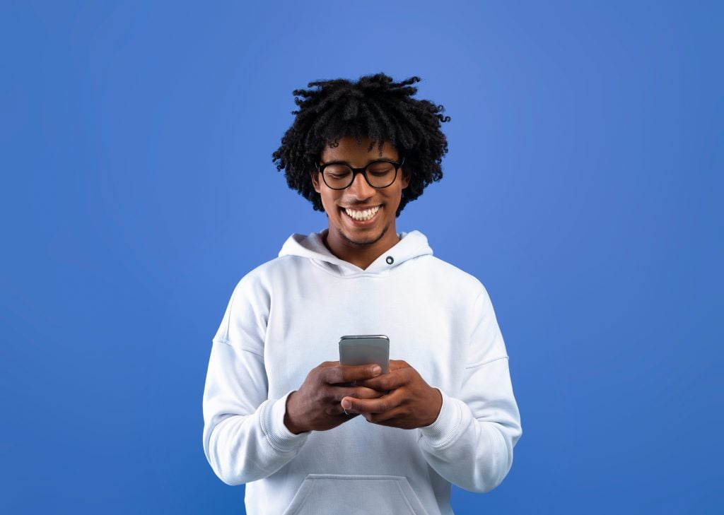 Happy black teenager looking at mobile phone screen, using new cool application on blue studio background