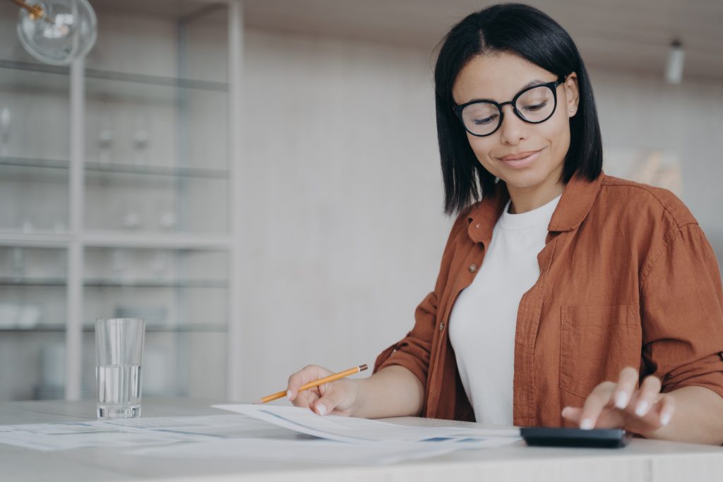 Female employee in glasses counts expenses on calculator, planning financial project budget at desk