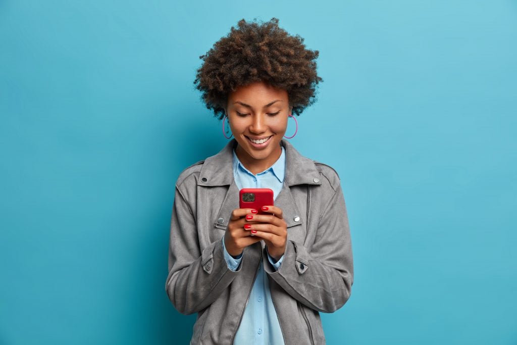 Happy smiling curly haired young woman types message on mobile phone, looks with glad expression at display, wears grey jacket, isolated on blue background. People, lifestyle, technology concept