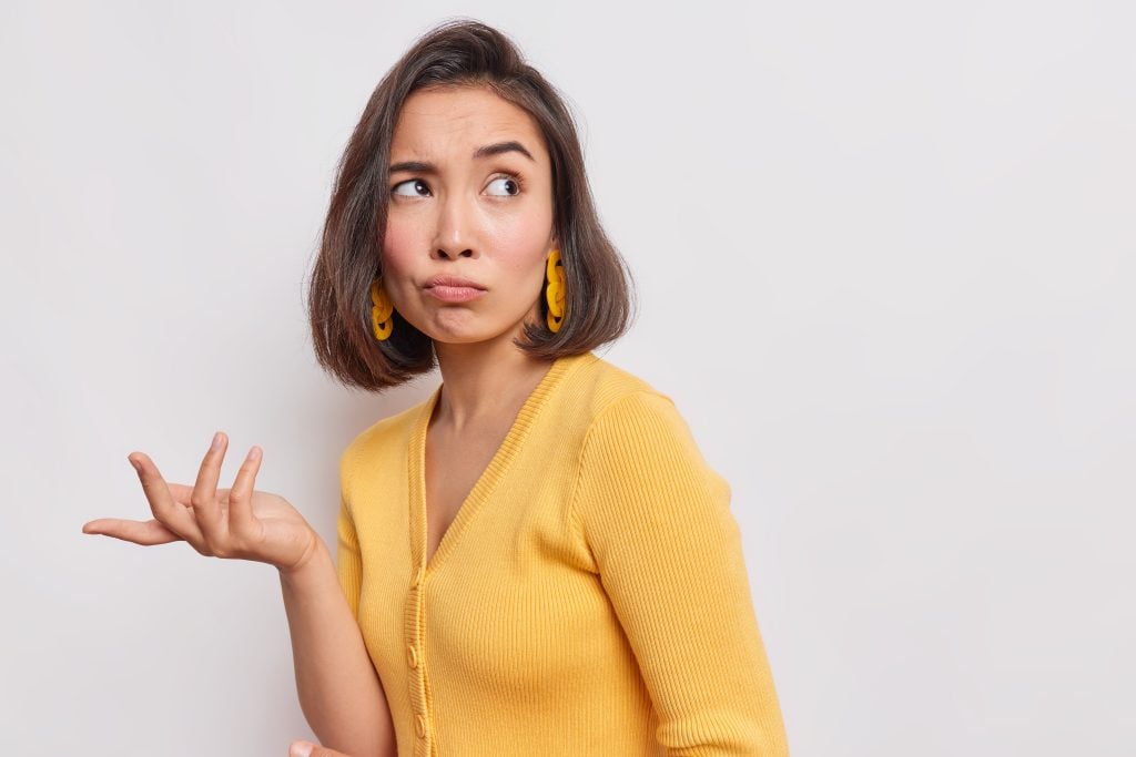 Indignant displeased Asian woman concentrated away has sad expression raises hand with clueless look wears yellow jumper earrings poses against white background copy space for text or promotion
