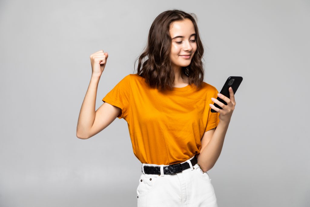 Portrait of a satisfied young woman holding mobile phone and celebrating isolated over gray background