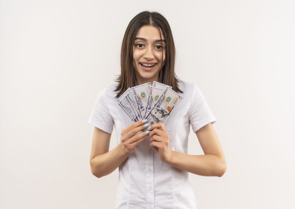 young girl in white shirt showing cash smiling cheerfully with happy face standing over white background