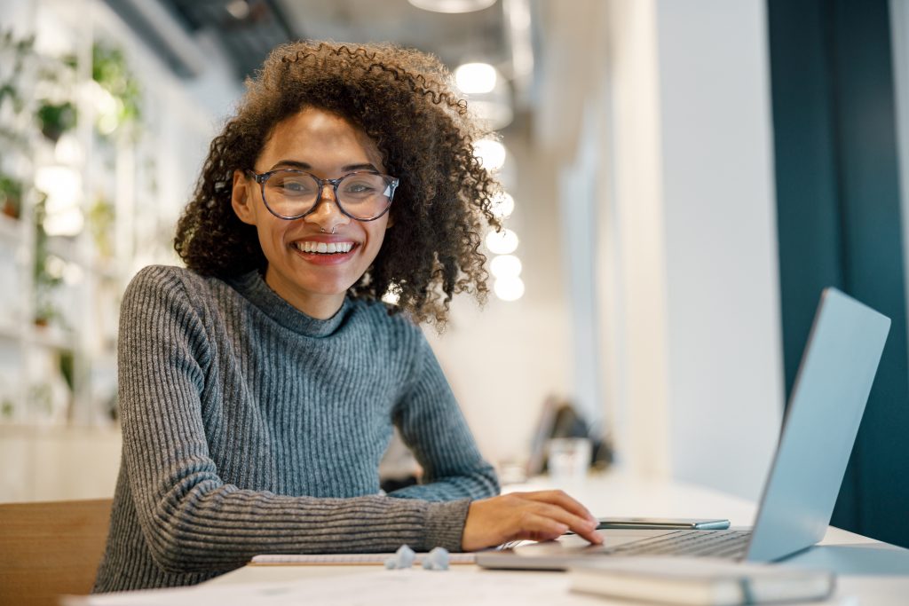 Smiling african woman freelancer working laptop while sitting in coworking and looking at camera