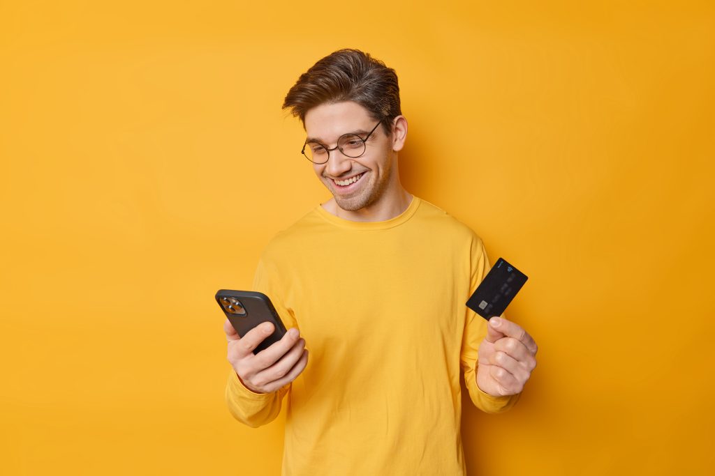 Cheerful guy with dark hair uses mobile phone and banking card for making shopping online orders something in online store enters information does secure payment isolated over yellow background