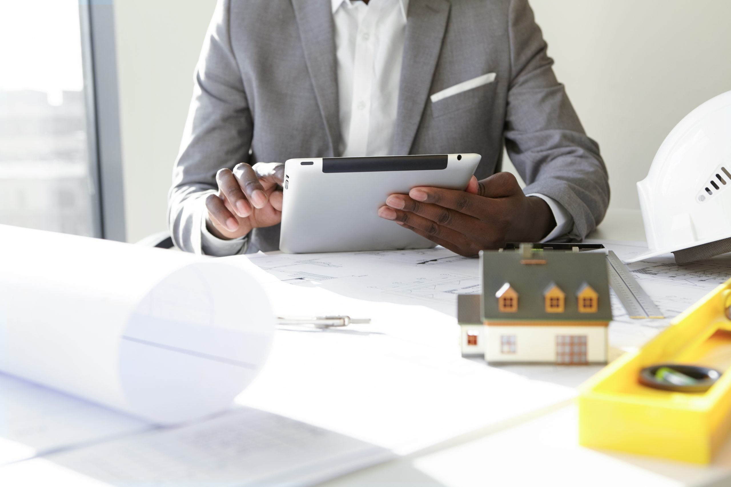 Selective focus. Design and architecture. Cropped shot of African architect holding digital tablet, developing new real estate project, sitting at desk with architectural tools and scale model house
