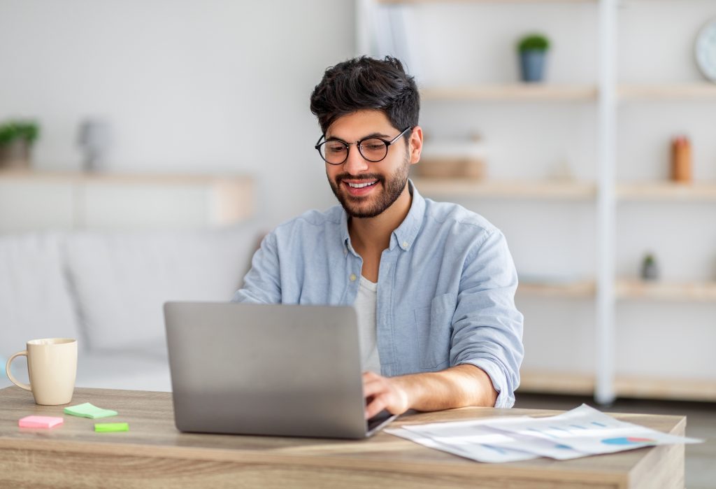 Portrait of happy arab freelancer man sitting at desk with laptop computer at home office, looking and smiling at screen