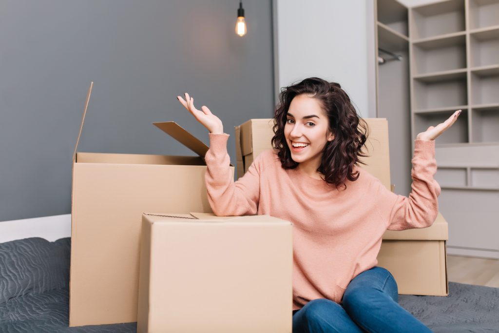 Excited young woman on bed surround boxes, carton smiling to camera in modern apartment. Moving to new flat, expressing true positive emotions at new home with modern interior.