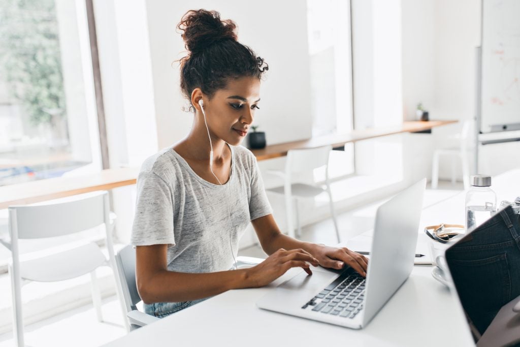 Romantic african girl with trendy hairstyle sitting at her workplace and analysing data. Indoor portrait of black female student working with laptop before exam..