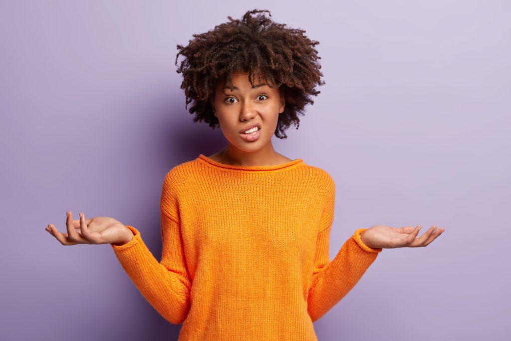 Isolated shot of confused beautiful woman with dark skin, curly haircut, spreads hands sideways, smirks face, feels doubt while makes choice, dressed in casual jumper, isolated over purple wall