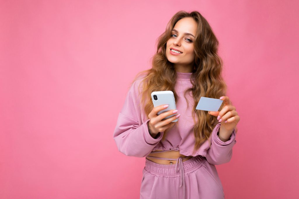 Photo of attractive positive smiling young blonde curly female peson wearing pink clothes isolated over pink background using mobile phone making payment using credit card looking at camera
