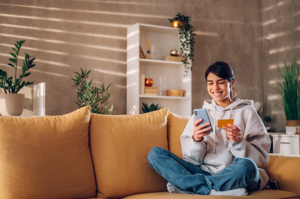 Beautiful smiling woman using smartphone and shopping online with credit card while sitting on a sofa when relaxing in a living room at home. Internet banking app, paying rent and bills online.
