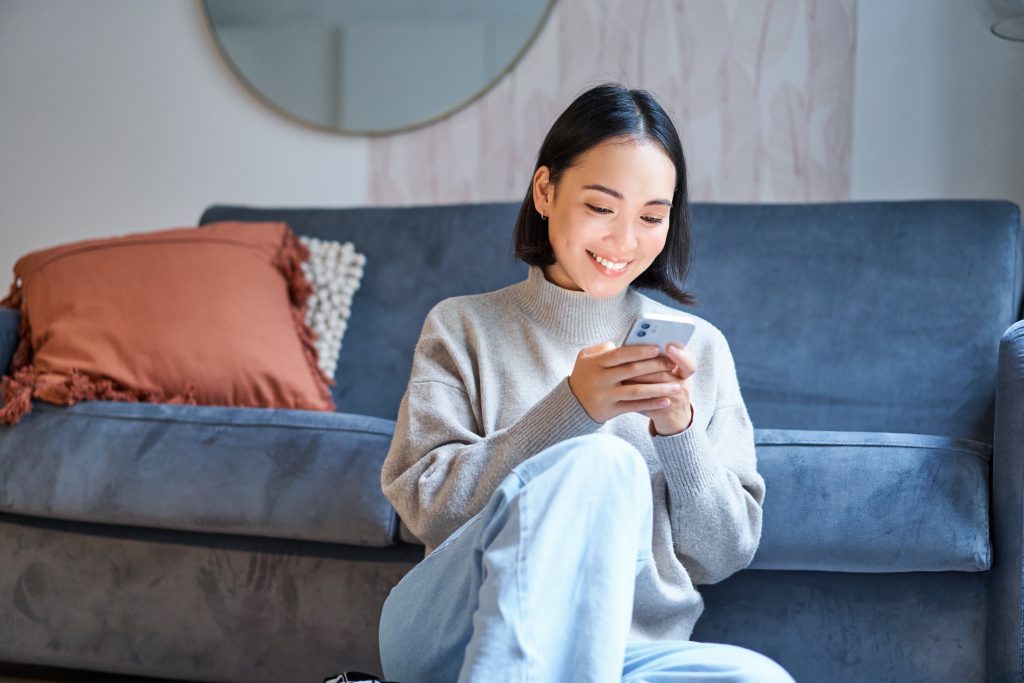 People and technology. Smiling asian woman sitting at home, using her mobile phone, typing message, browing internet or shopping online from smartphone app