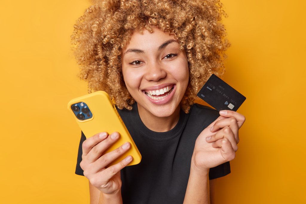 Happy curly female shopper enjoys easy paying for goods online smiles broadly shows white teeth uses mobile phone and credit card dressed in casual black t shirt isolated over yellow background