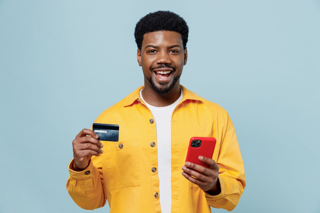 Young fun man of African American ethnicity in yellow shirt using mobile cell phone hold credit bank card do online shopping order delivery booking tour isolated on plain pastel light blue background.